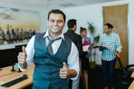 Smiling businessman showing thumbs up in a lively office environment with colleagues in the background.